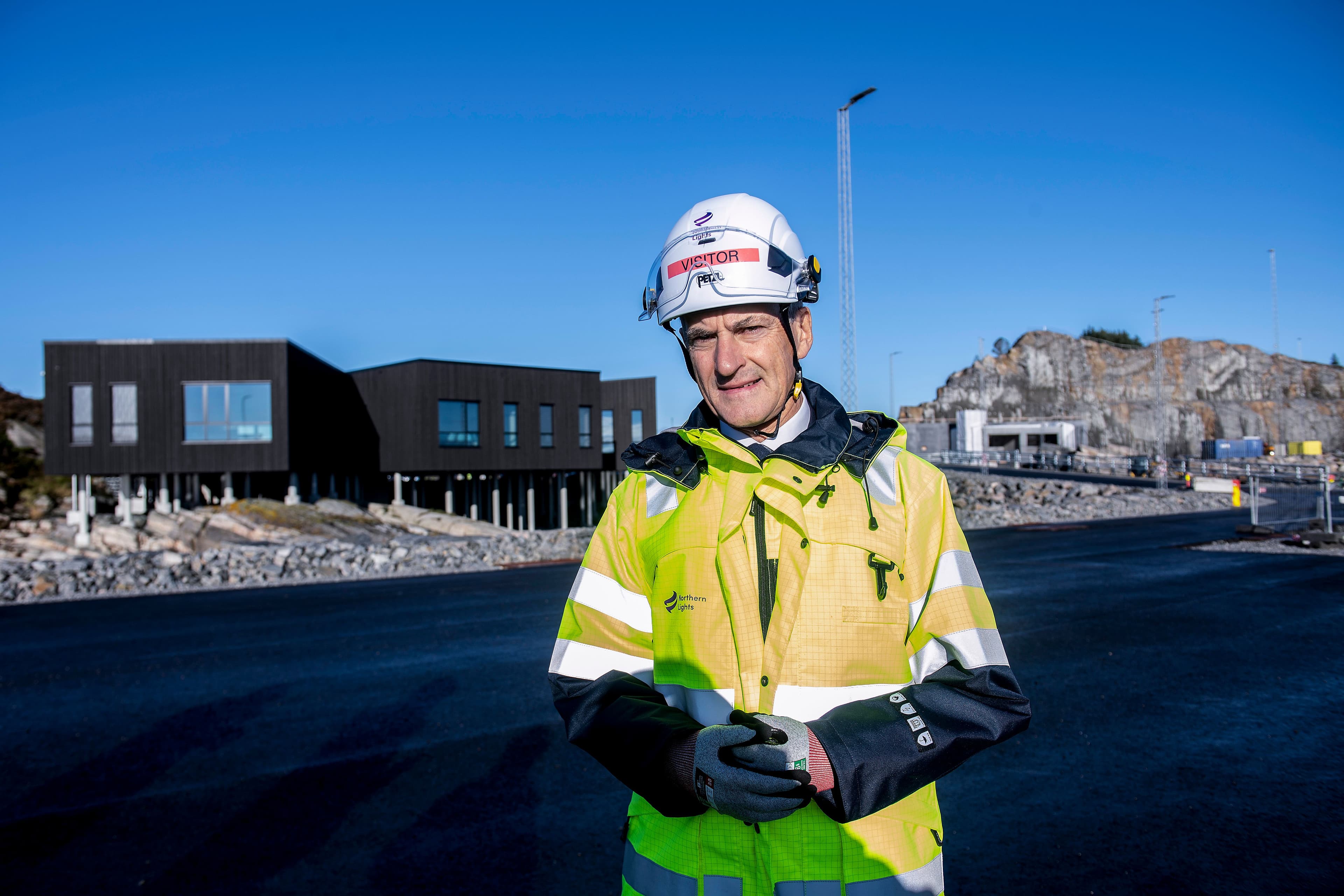 En mann med hjelm og refleksjakke står utendørs, med moderne bygninger og en steinete åsside i bakgrunnen.