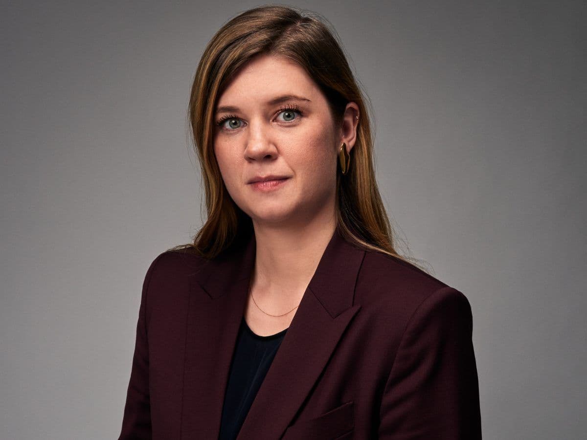 En kvinne med langt brunt hår iført en mørk blazer og svart topp poserer mot en vanlig, grå bakgrunn.