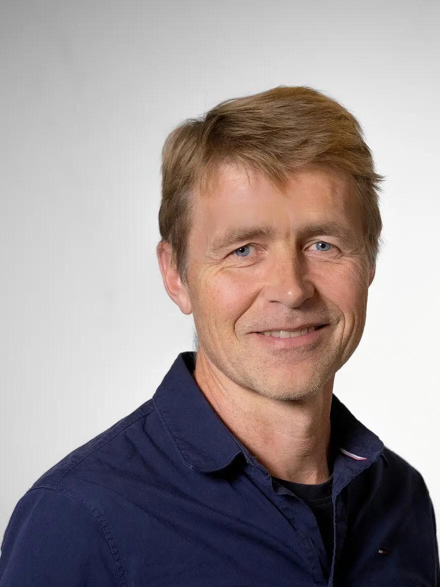 En middelaldrende mann med lysebrunt hår, iført en mørkeblå skjorte med krage, smiler til kameraet mot en enkel lys bakgrunn.