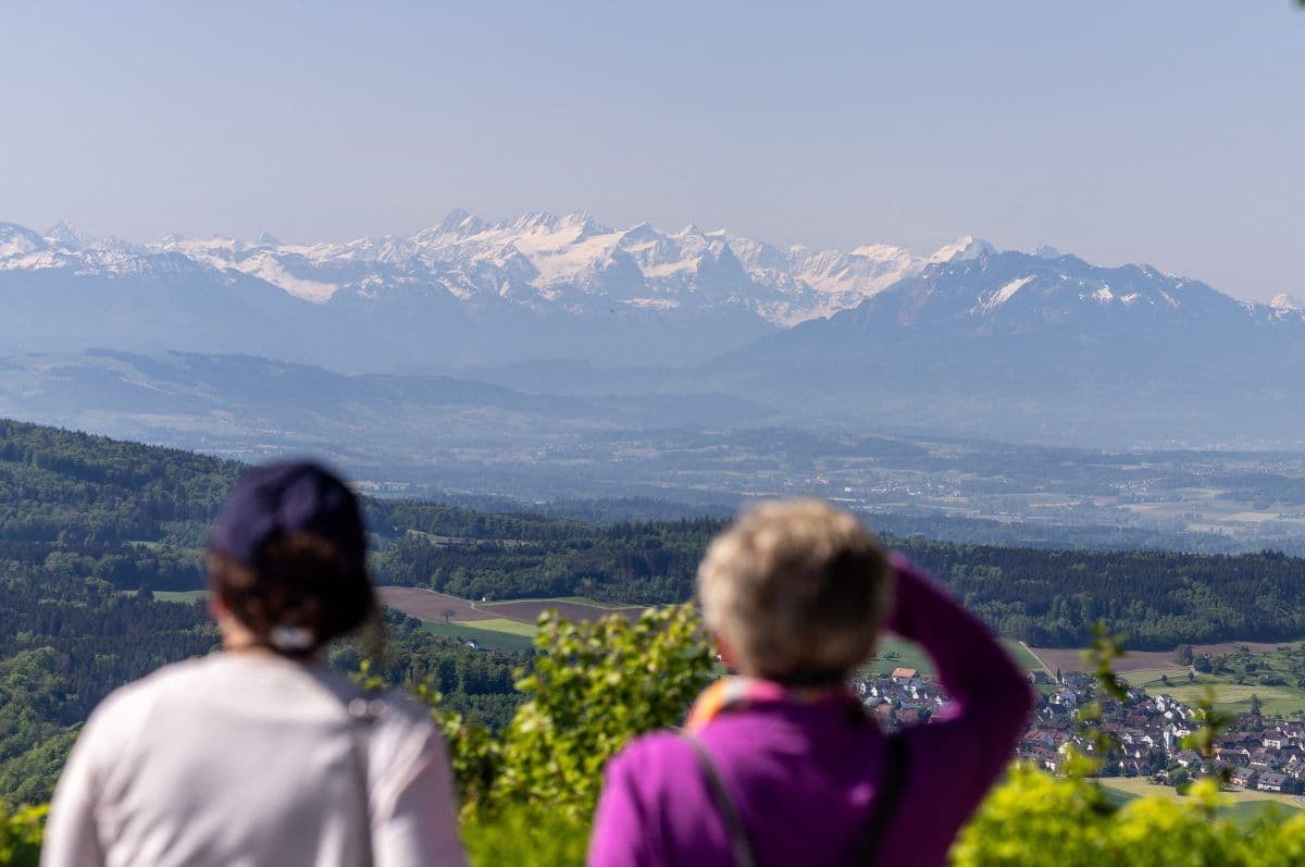 To personer ser på et naturskjønt landskap med fjerne snødekte fjell, grønne åser og en by nedenfor under en klar himmel.