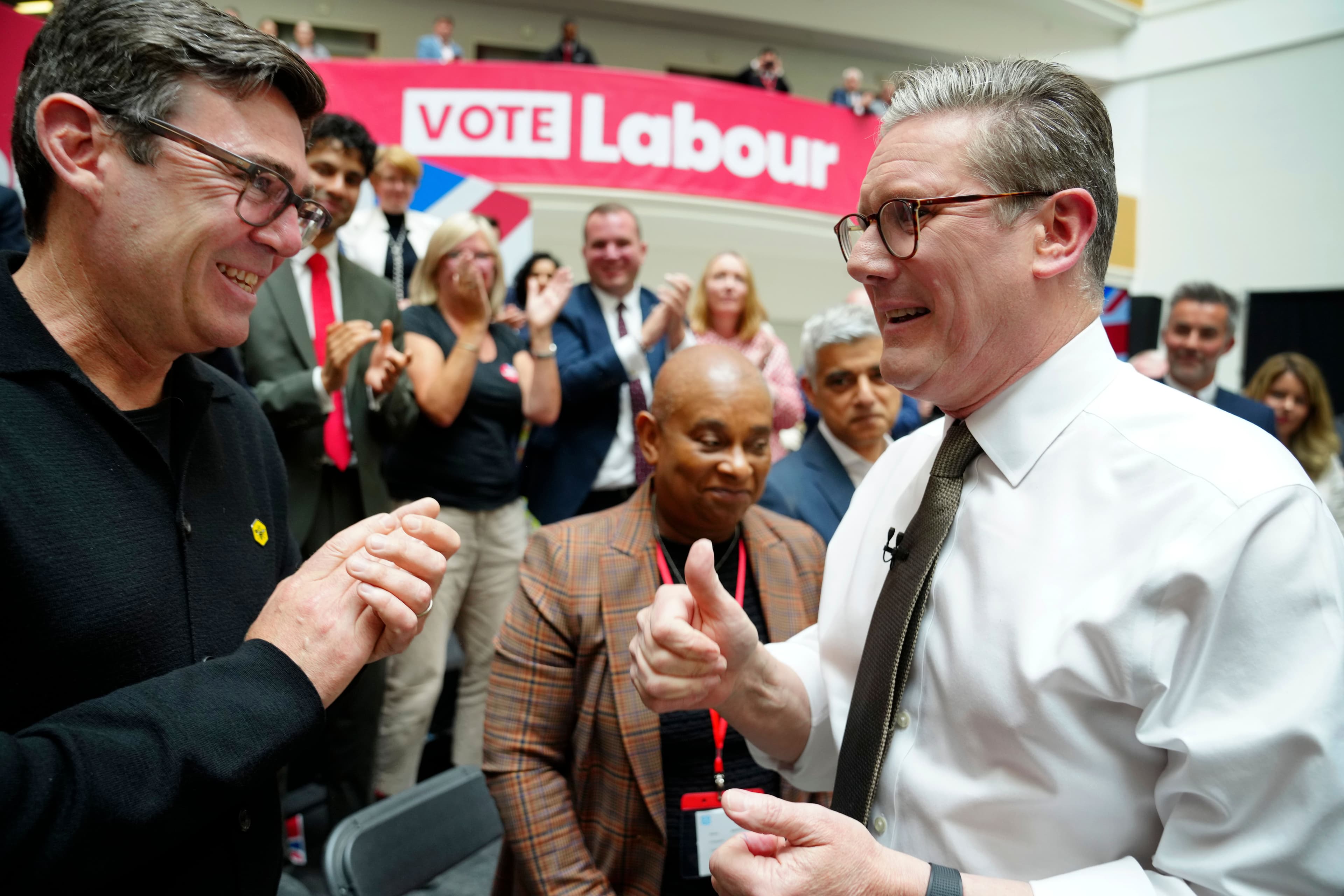 Andy Burnham smiler mens Keir Starmer viser tommelen opp på et Labour-valgmøte, med et «STEM Labour»-banner og en jublende folkemengde bak dem.