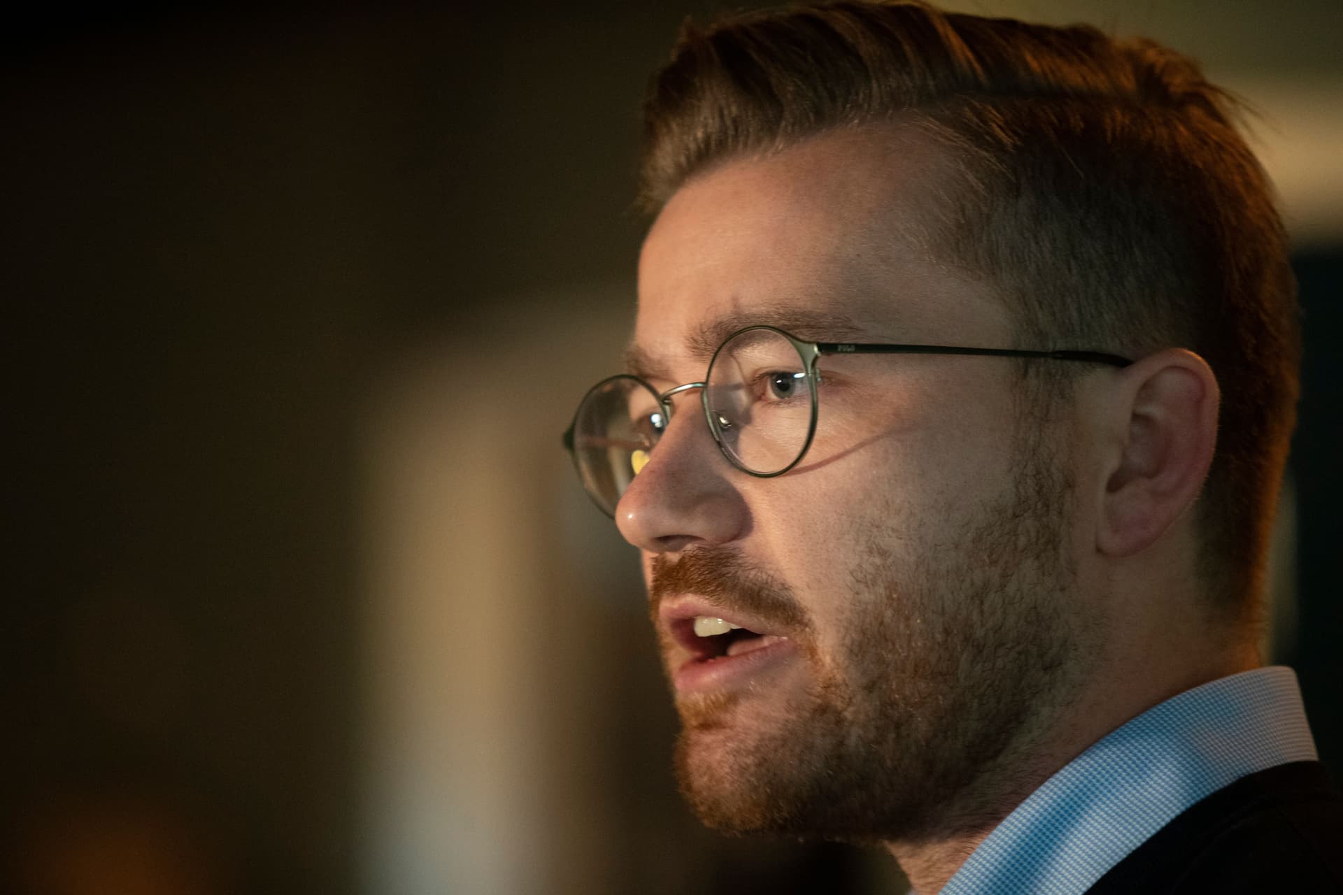 A man with a light brown beard and round glasses speaks, looking to the right.