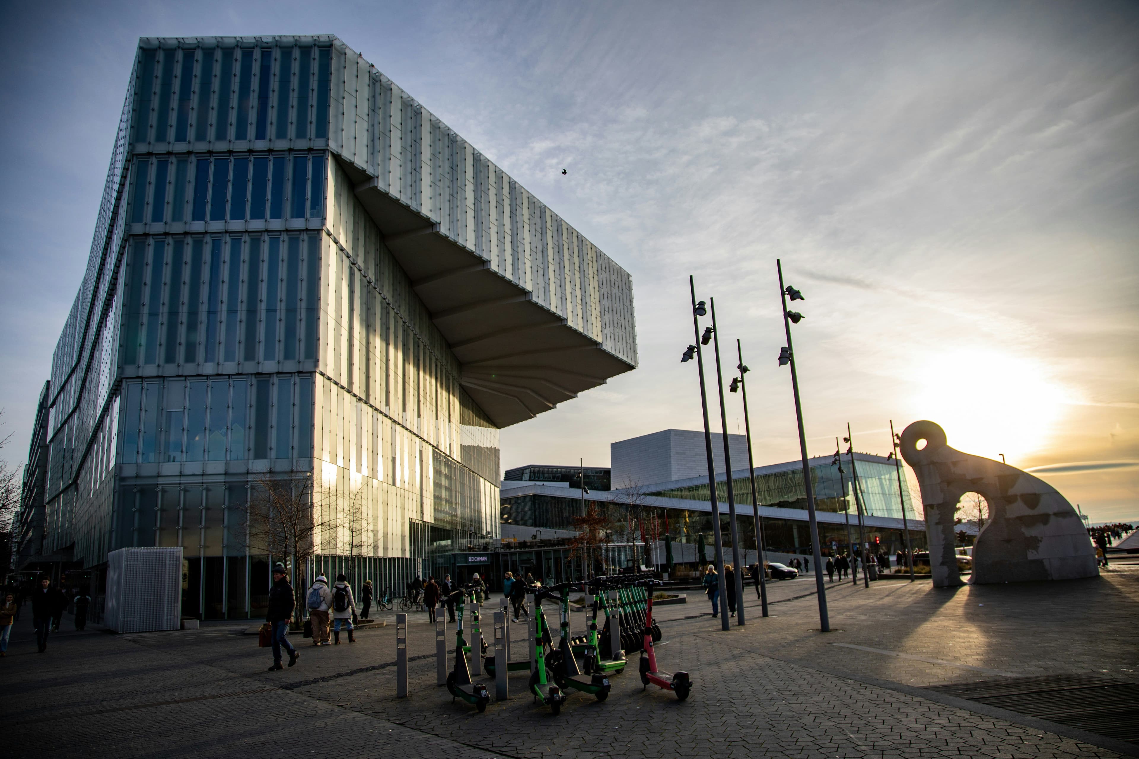 Et moderne glassbygg, offentlig plass med mennesker, sparkesykler og en abstrakt skulptur under en solnedgangshimmel.