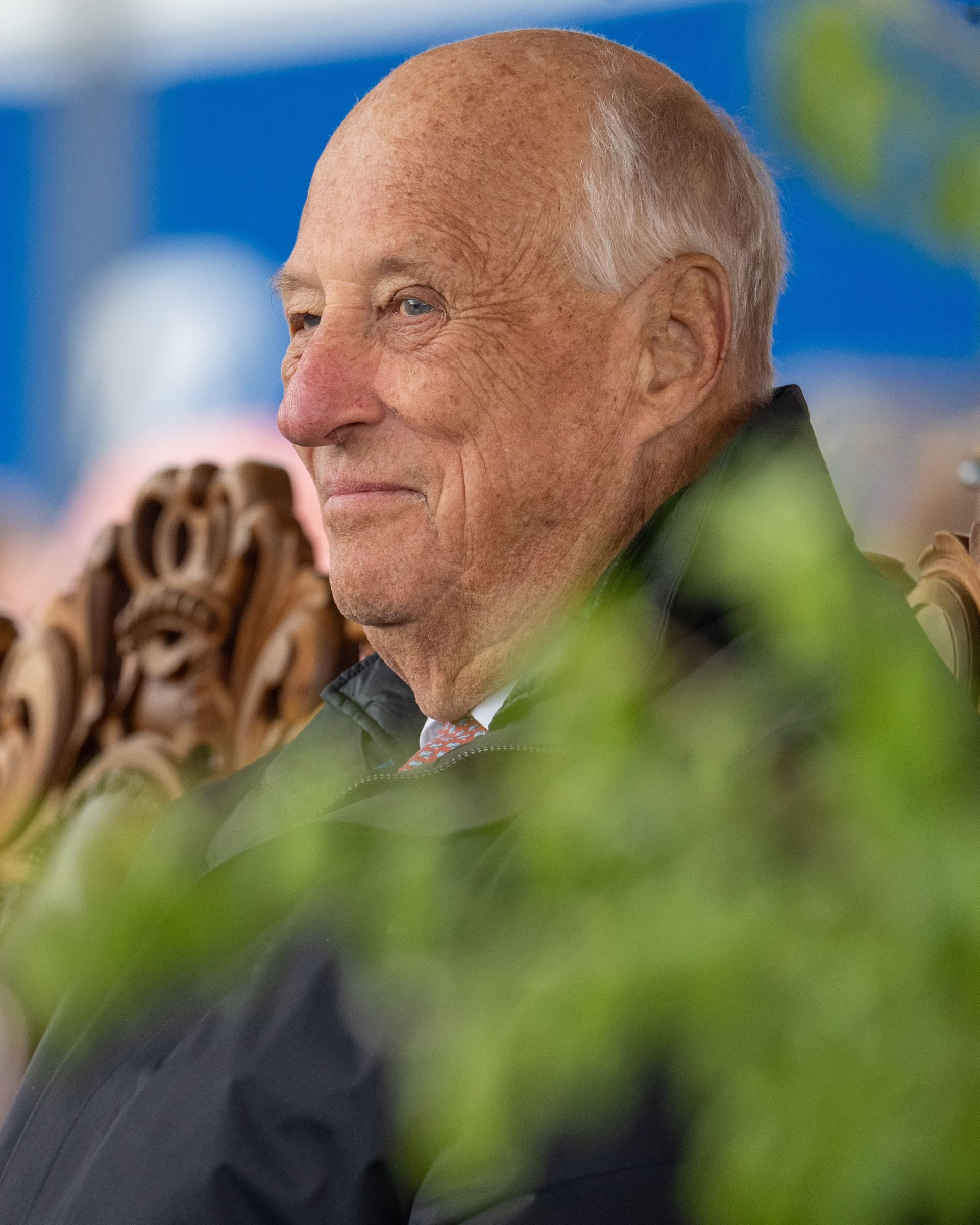A smiling older man with white hair and a dark jacket, partly obscured by green foliage.