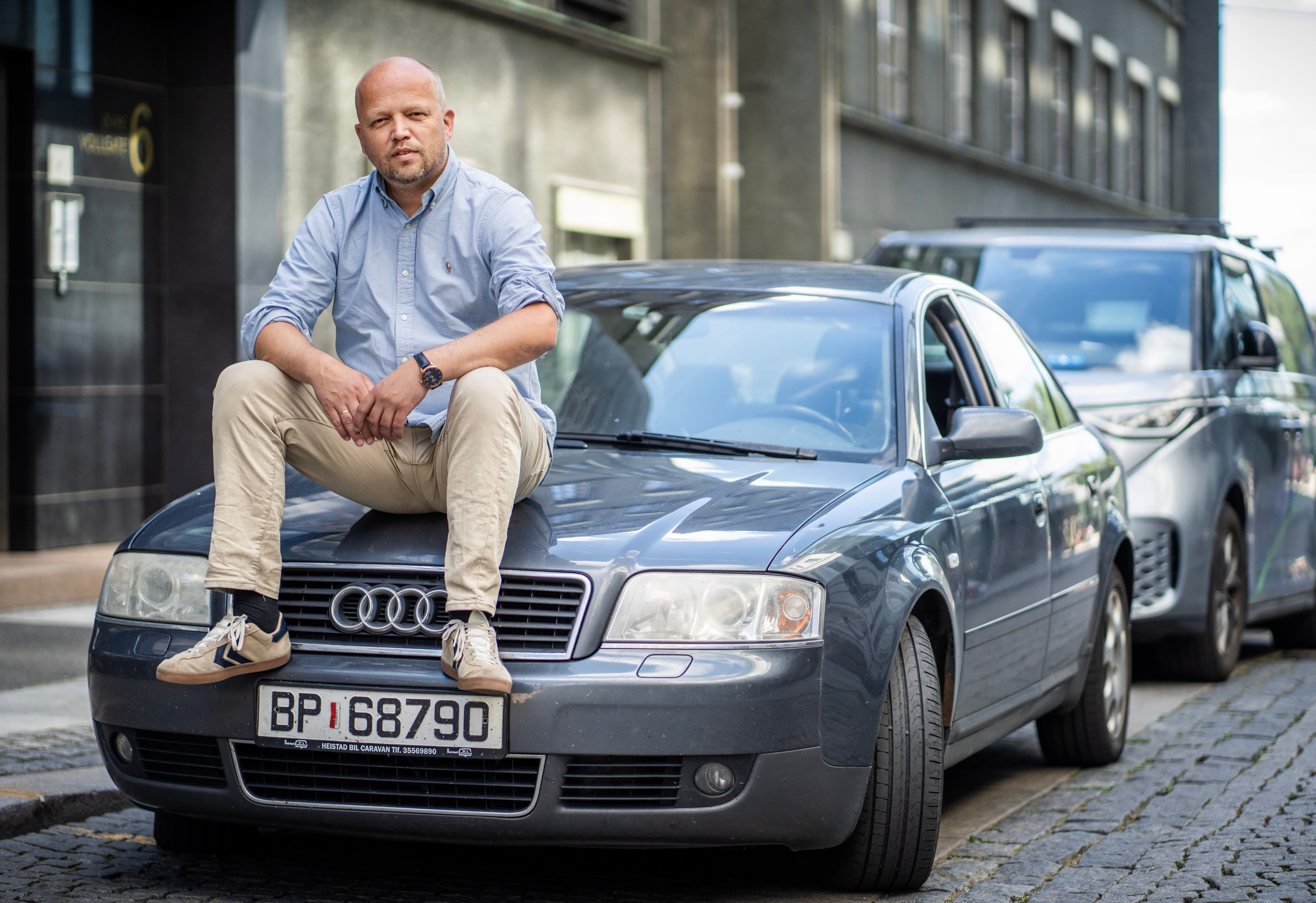 En skallet mann sitter på panseret til en grå Audi på en brosteinsbelagt gate.